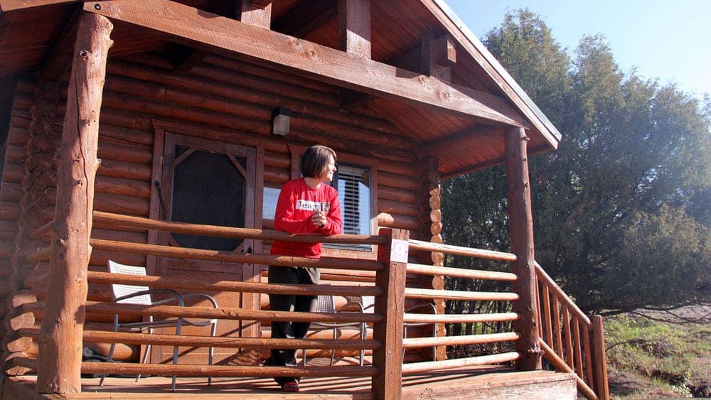 A person stands on the porch of a wooden cabin, looking out at the scenery.