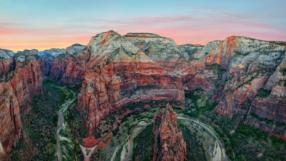 A panoramic view of rugged red rock formations and a winding river at sunset in Zion National Park.