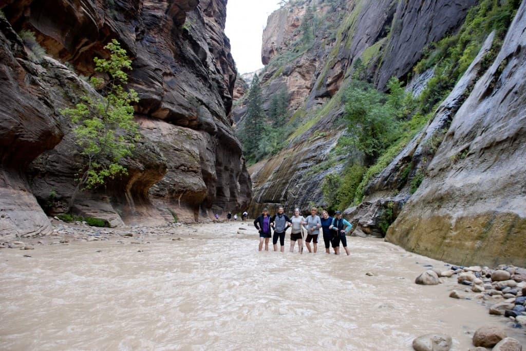 A group of six people wades through a shallow river in a narrow canyon surrounded by steep rock walls and vegetation.