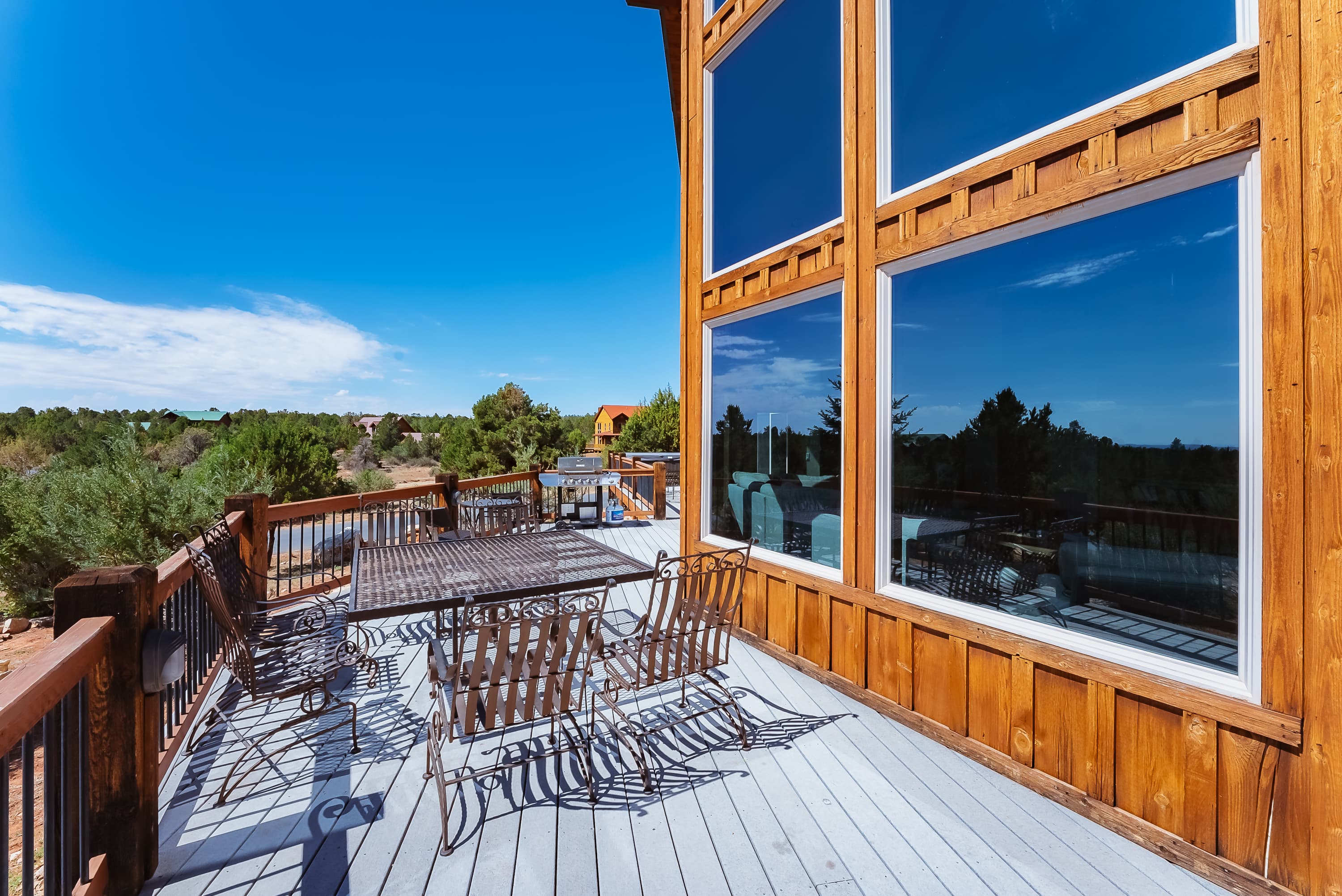 A wooden deck with metal furniture and large windows overlooking a blue sky and greenery.