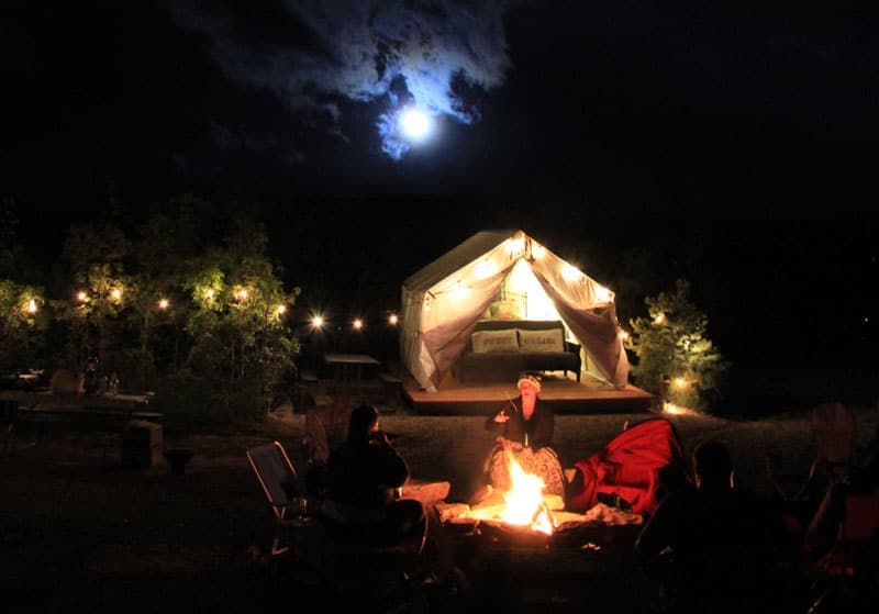 A group of people gathers around a campfire at night, with a lit tent and a bright moon in the background.
