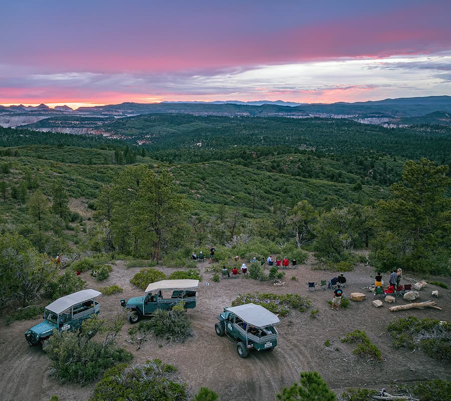 A panoramic view of a sunset over a forested landscape with several off-road vehicles and people gathered around a campsite.