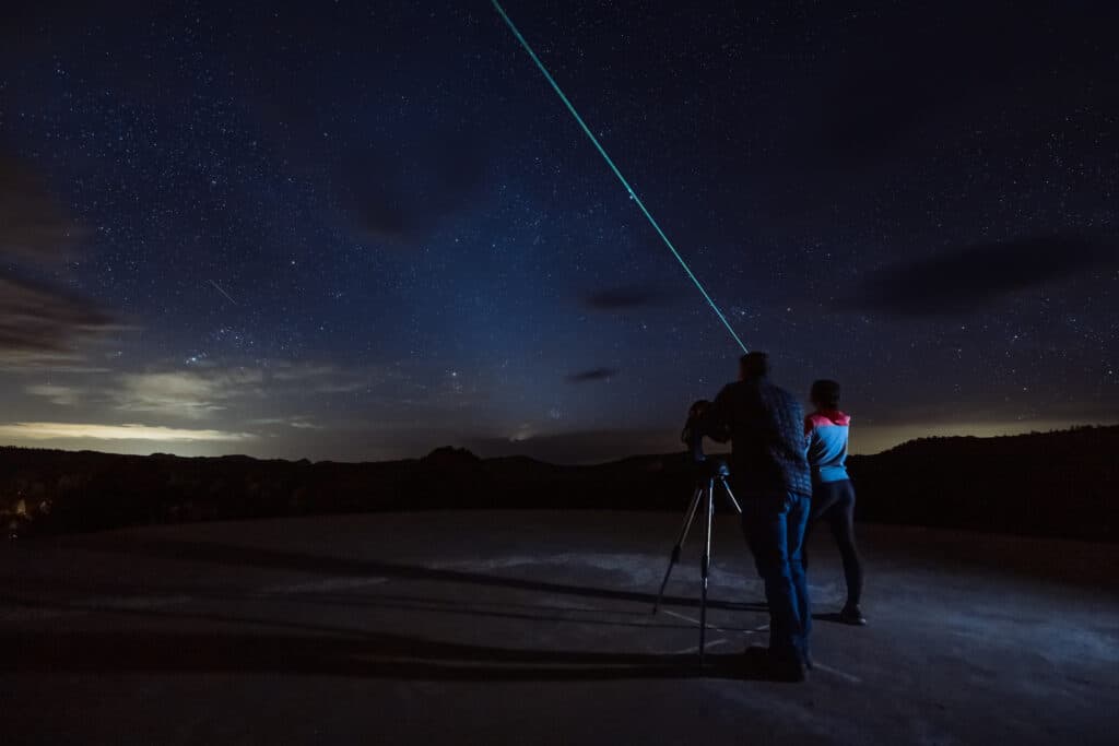 Two people on a rooftop observe the night sky, pointing a laser at the stars.