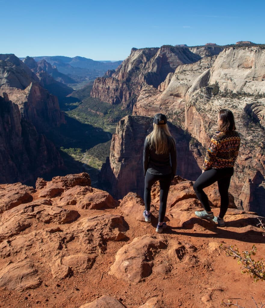 Two women stand on a rocky cliff overlooking a vast canyon landscape.