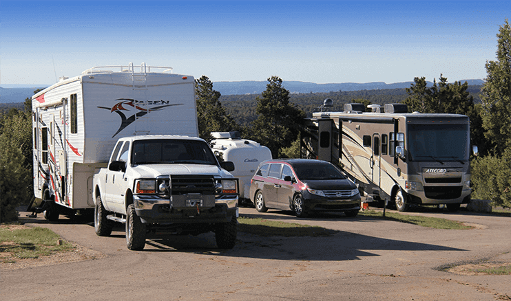 A white RV, a truck, and a silver car are parked at a scenic campsite surrounded by trees.