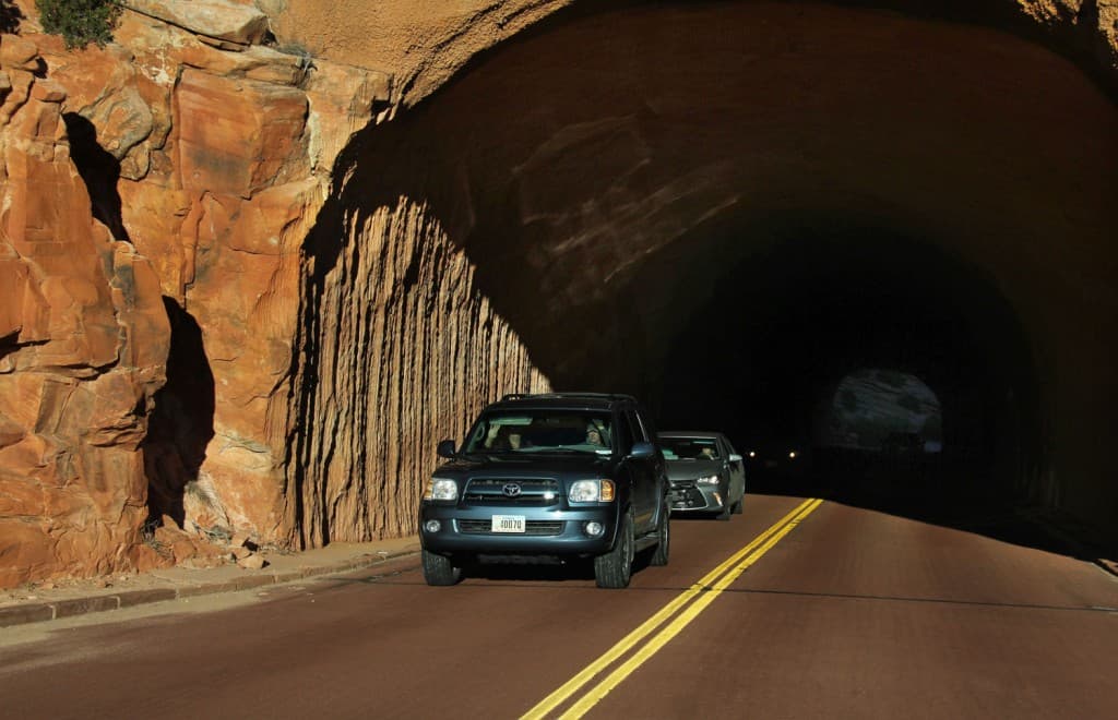 Two cars drive through a rocky tunnel on a sunlit road.