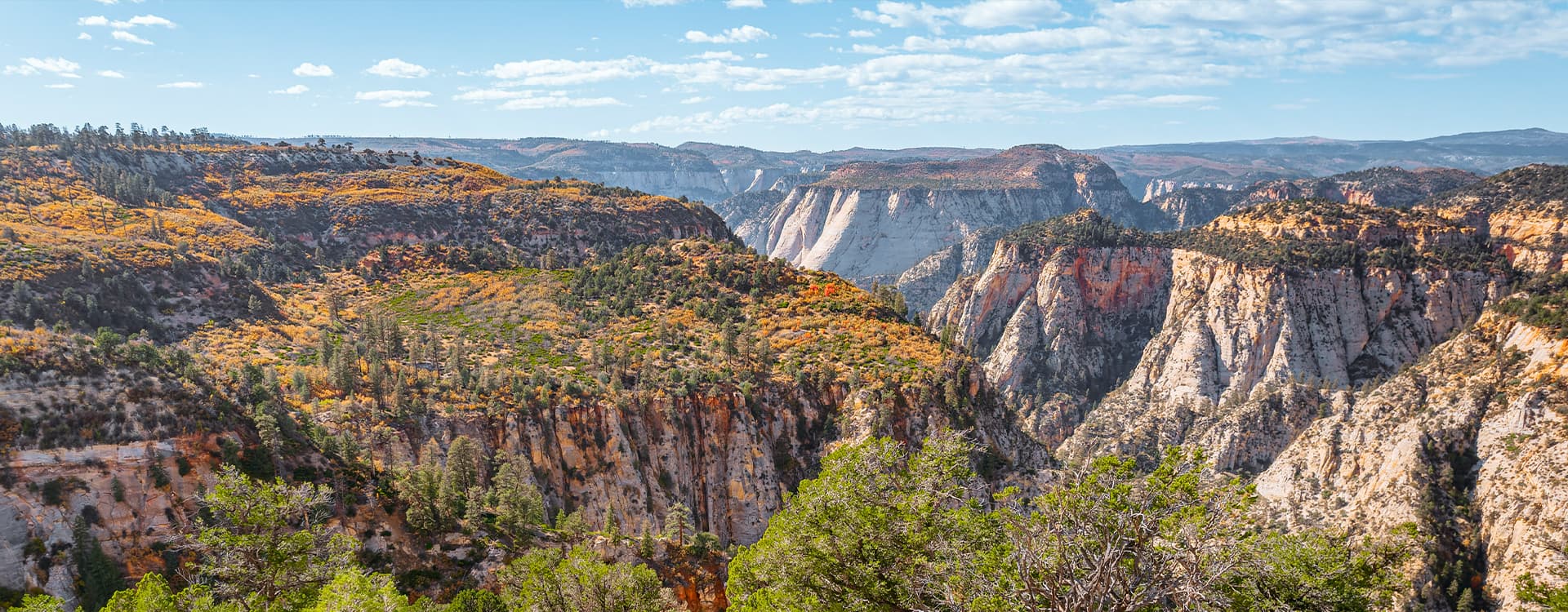A panoramic view of colorful canyon cliffs under a blue sky with scattered clouds.