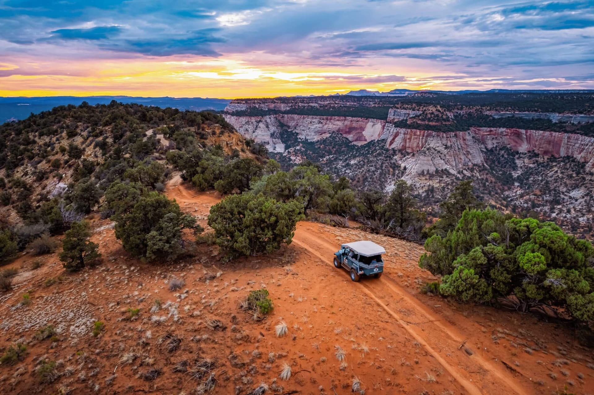 A Jeep navigates a dirt road surrounded by rugged terrain under a colorful sunset.