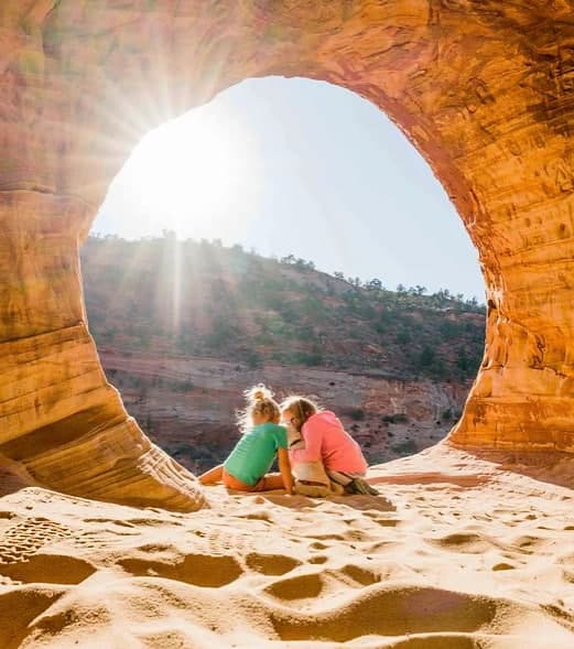 Two children sit on sandy ground inside a rock arch, bathed in sunlight.
