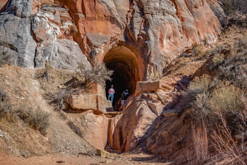 Two hikers stand near the entrance of a rocky tunnel in a desert landscape.