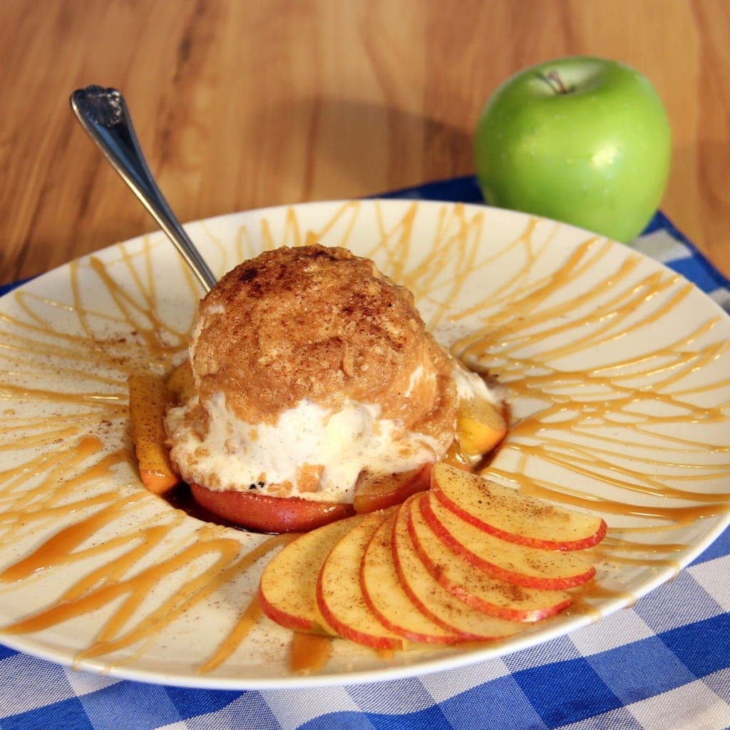 A dish featuring a scoop of ice cream topped with a crumble, served on apple slices, drizzled with caramel, next to a green apple.