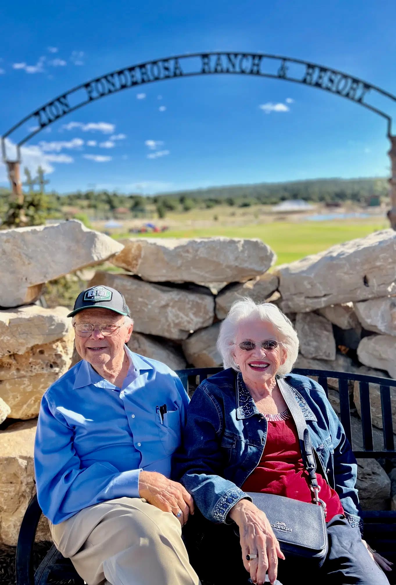 An elderly couple sits together on a bench in front of a stone archway at a ranch.