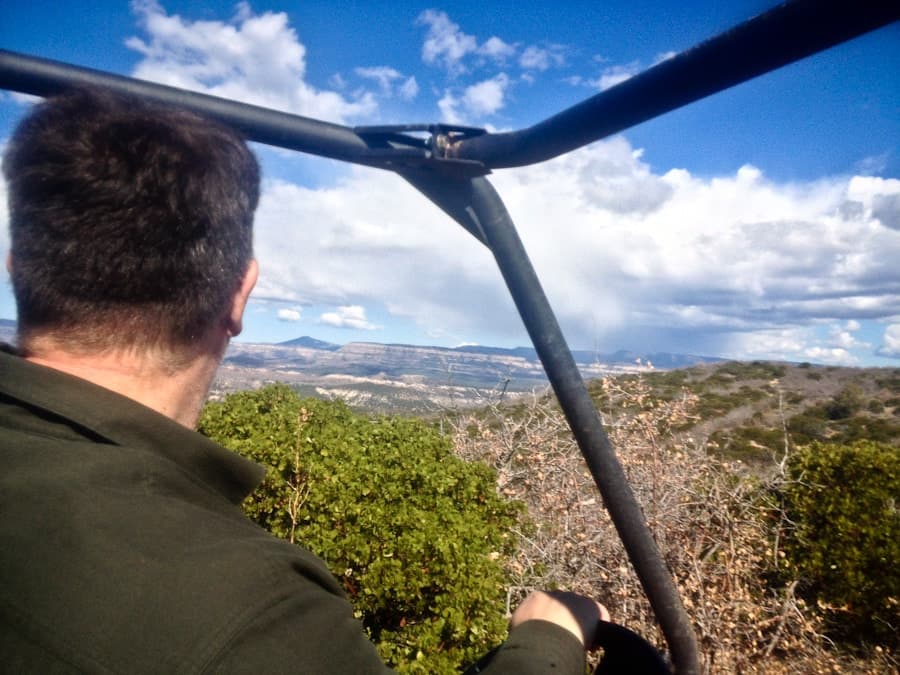 A person is seated in a vehicle, looking out over a scenic landscape with mountains and a blue sky.