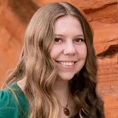 A smiling woman with long wavy hair stands in front of a red rock background.