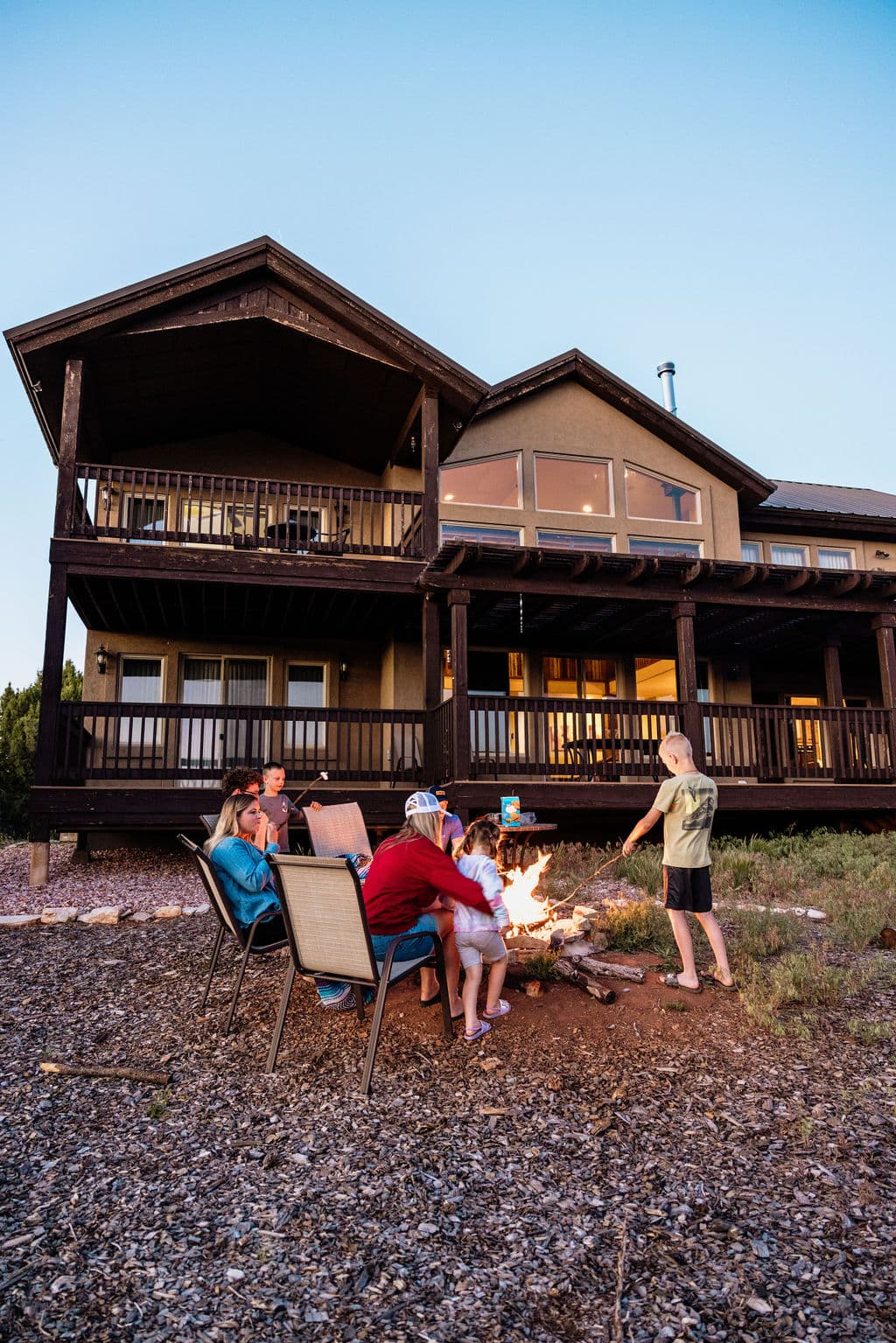 A group of people gather around a campfire in front of a large wooden house at sunset.