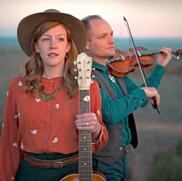 A woman with a guitar stands in front of a man playing the violin against a sunset backdrop.