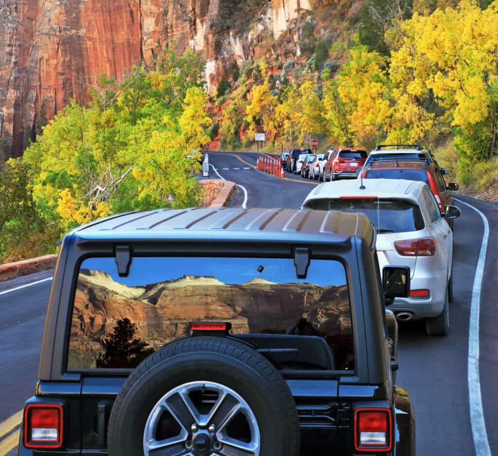 A line of cars is stopped on a scenic road with autumn trees and cliffs in the background.
