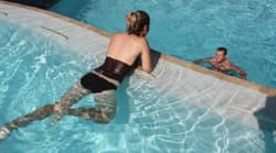 A woman in a swimsuit leans on the edge of a swimming pool while a man swims nearby.