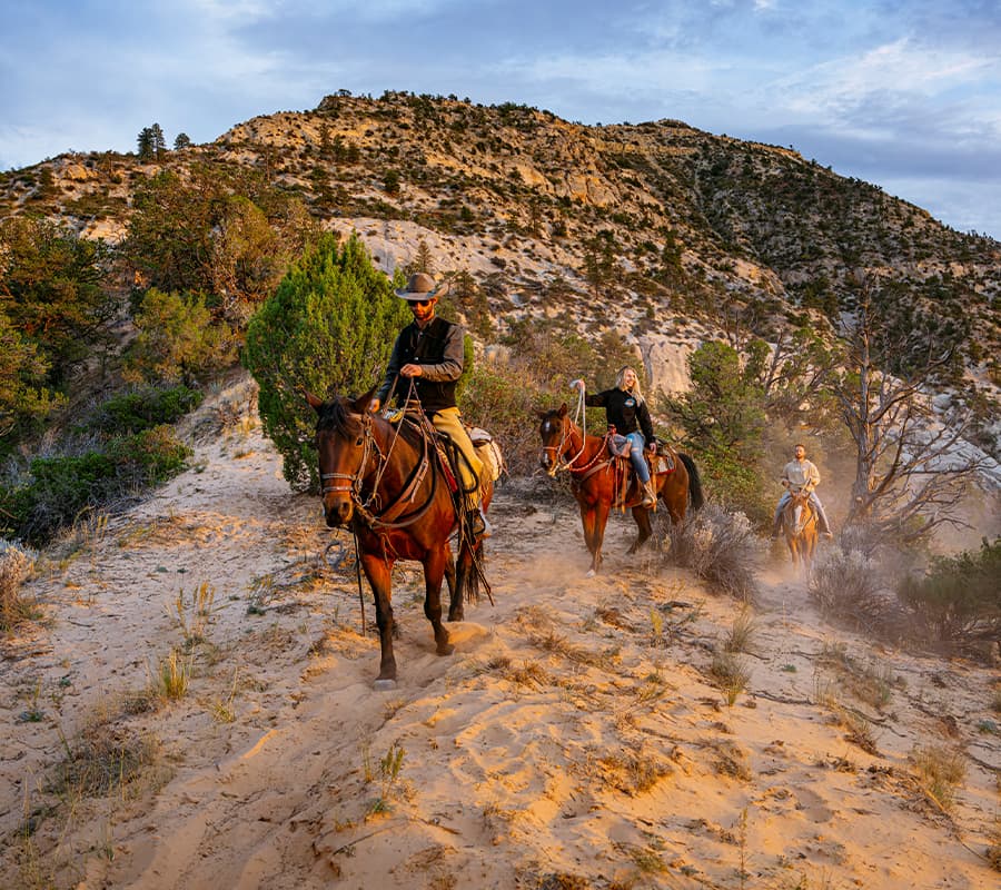 Three horseback riders navigate a sandy trail surrounded by rugged mountains and vegetation.
