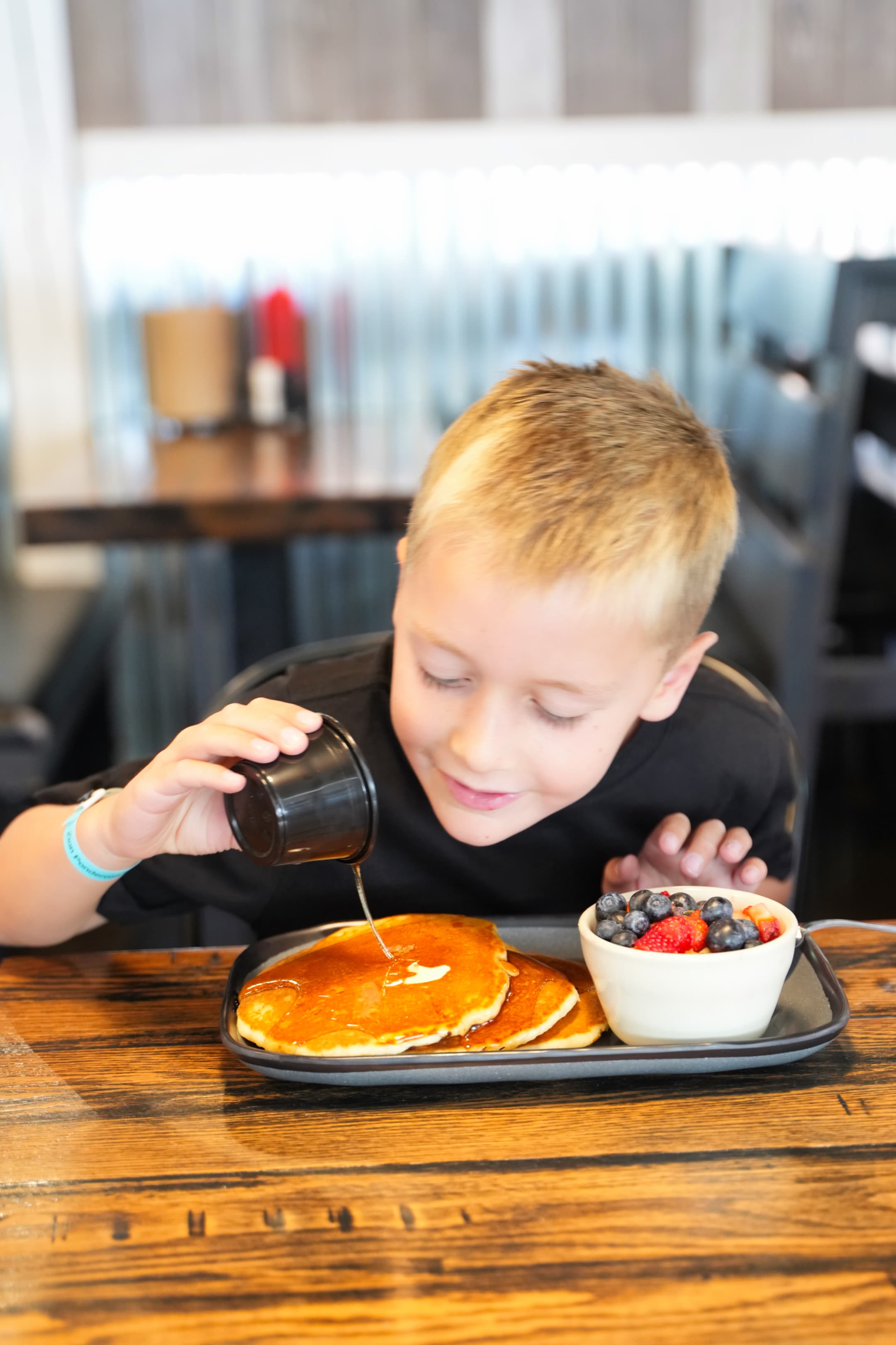 A young boy joyfully pours syrup over a stack of pancakes beside a bowl of fresh berries.
