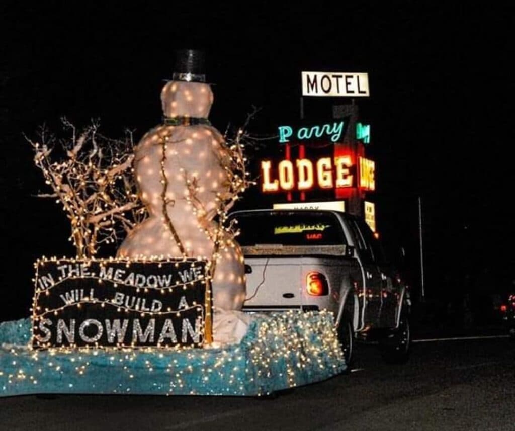 A decorated snowman with lights and a sign reading "In the meadow we will build a snowman" next to a truck in front of a brightly lit motel sign at night.