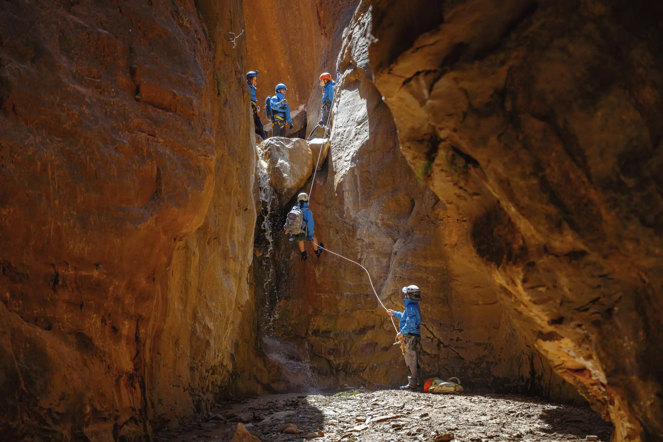 A group of climbers ascends a rocky canyon wall, navigating steep terrain under bright sunlight.