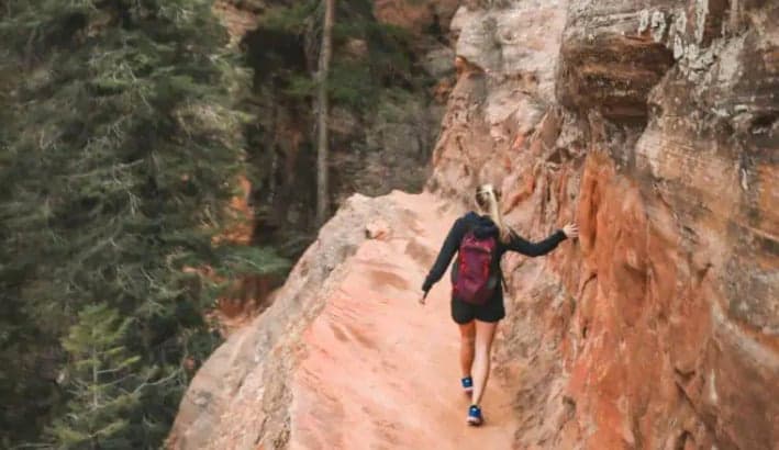 A hiker walks along a narrow trail beside a rocky cliff in a forested area.