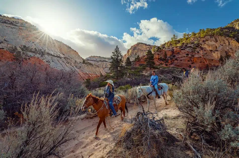 Three riders on horseback traverse a sandy trail through rugged canyon landscapes under a bright sky.