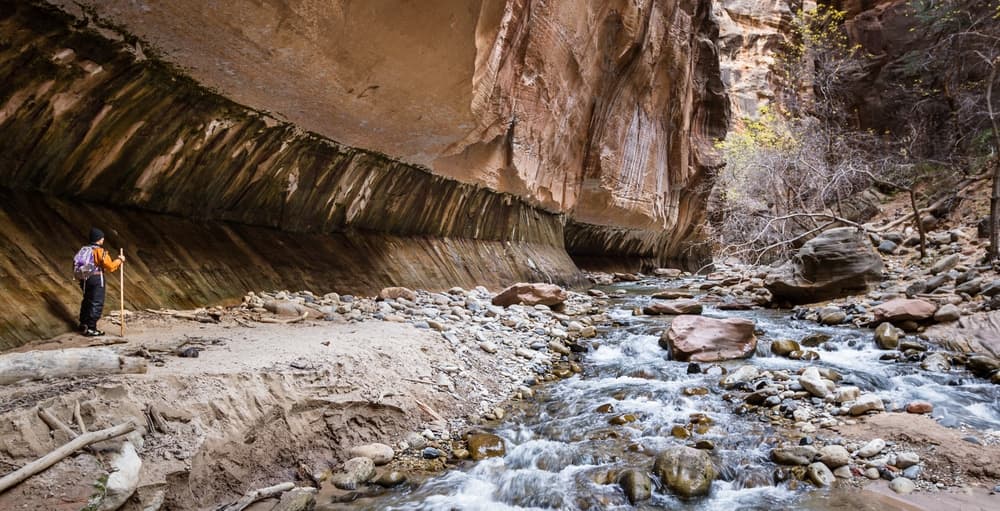 A hiker stands by a flowing stream in a narrow canyon with towering rock walls.
