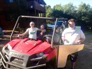 Three people pose with a red Polaris off-road vehicle in a wooded area.