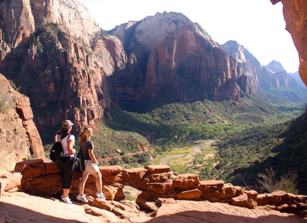 Two people stand on a rocky ledge overlooking a lush valley surrounded by towering red cliffs.