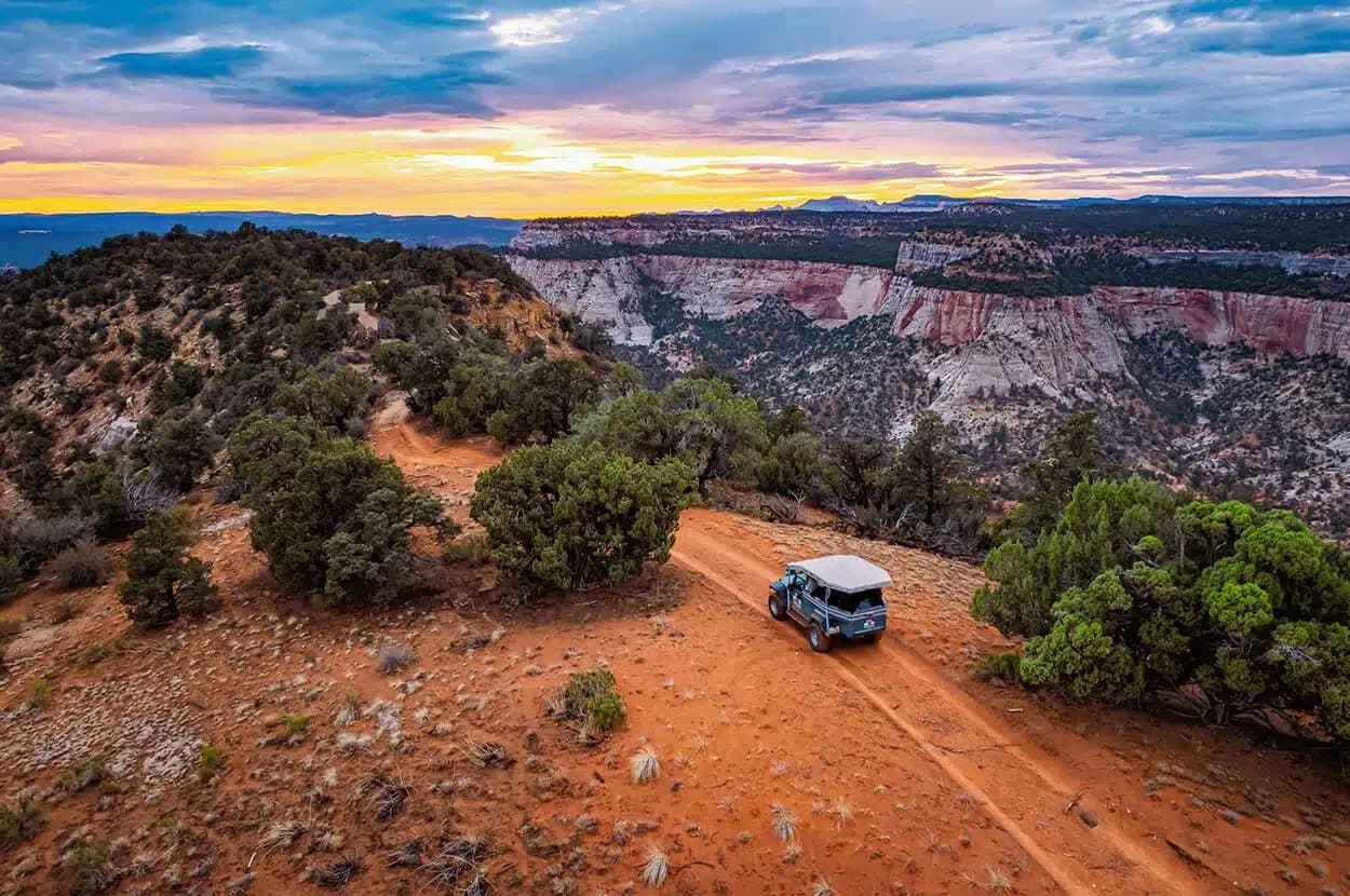 A vehicle parked on a red dirt road overlooking a scenic canyon at sunset.