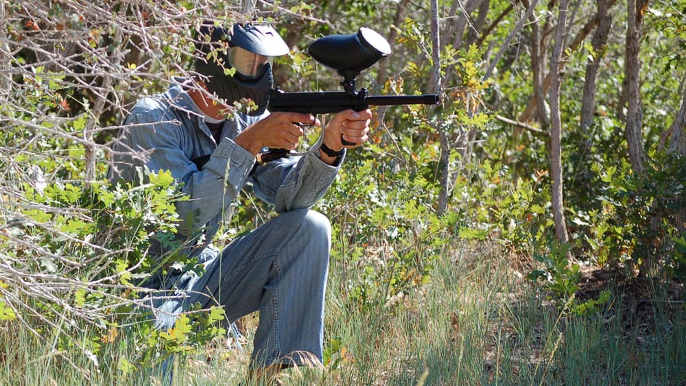 A person crouches in a wooded area, aiming a paintball gun.
