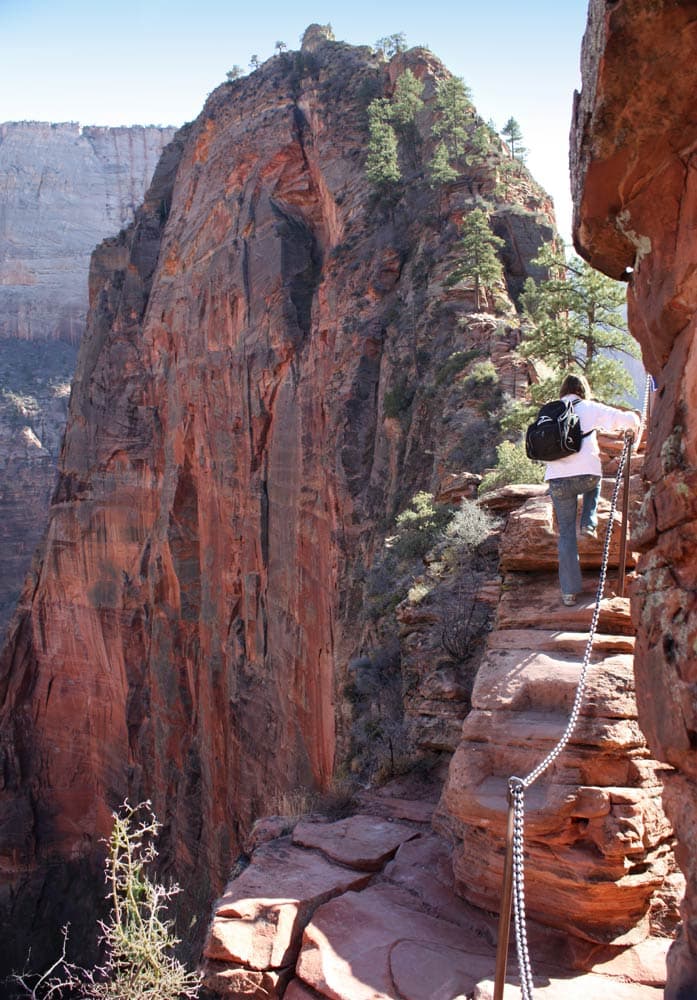A person navigates a narrow trail along a steep cliff with a chain for support.