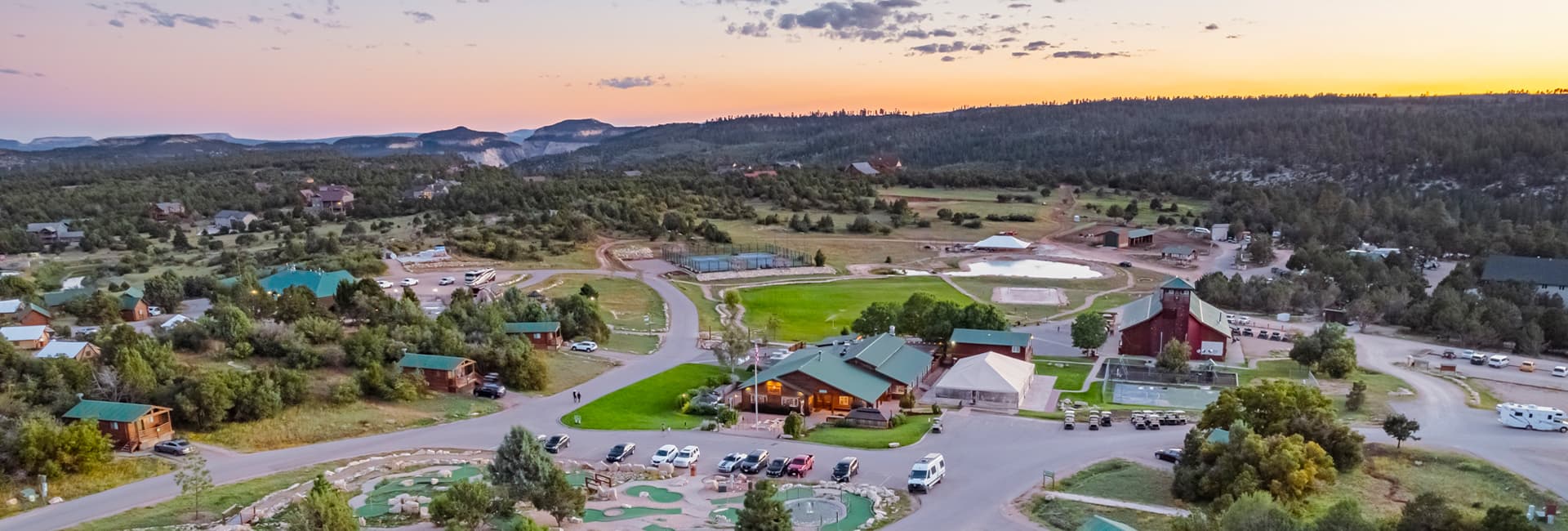Aerial view of a rustic lodge area surrounded by green fields and mountains at sunset.