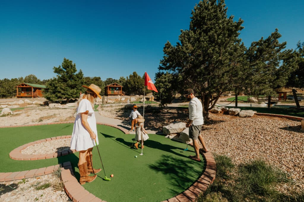 A family enjoys a game of mini-golf on a sunny day surrounded by trees and cabins.