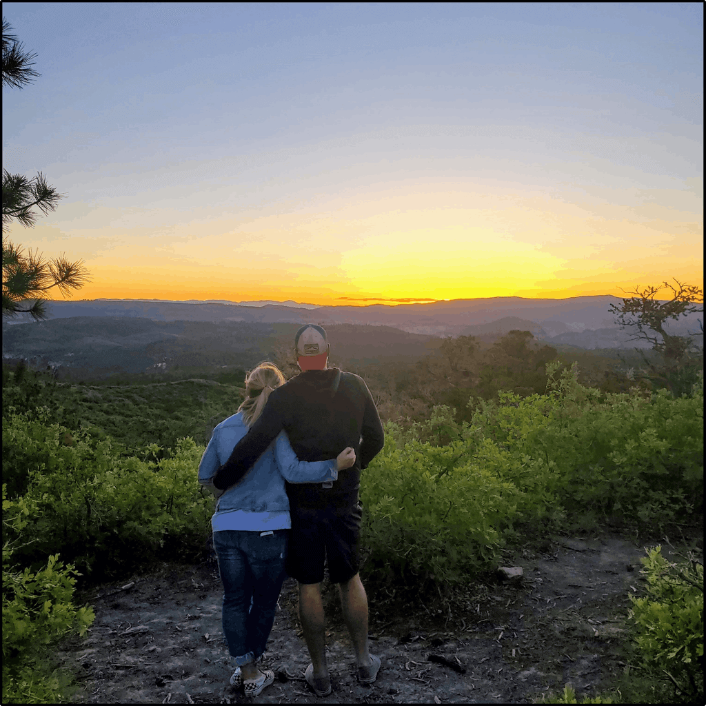A couple stands together, overlooking a vibrant sunset amidst hills and greenery.