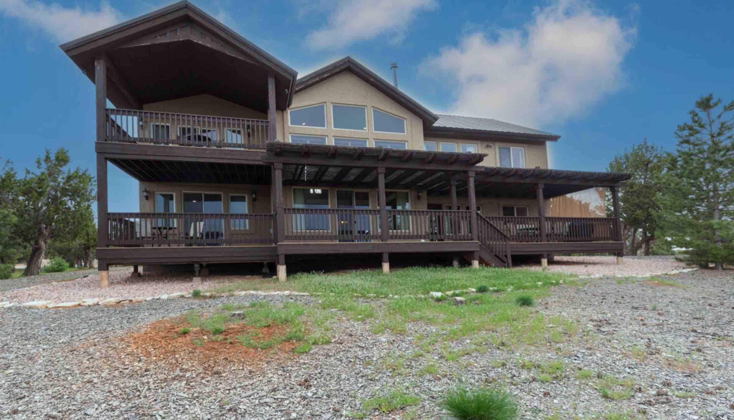 A large, two-story house with a wooden deck, surrounded by greenery and gravel.