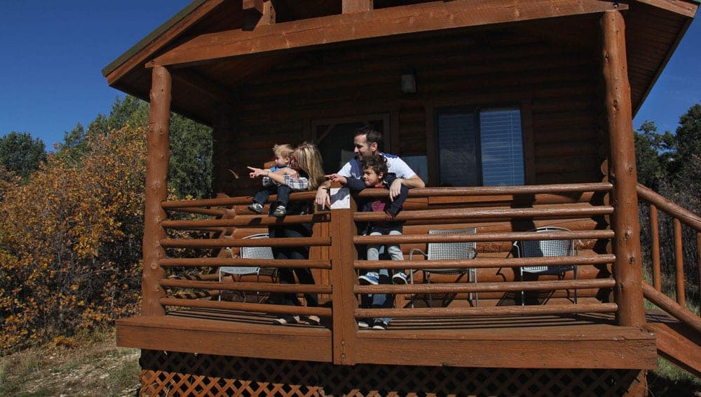 A family stands on the balcony of a log cabin, pointing and looking at the scenery.