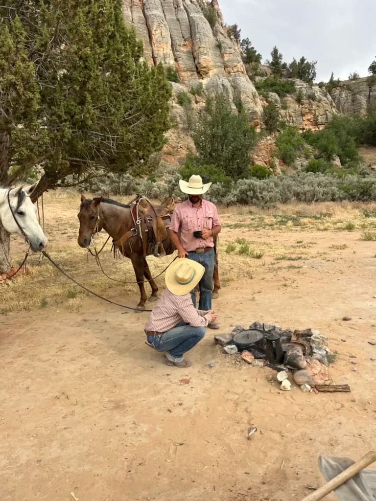 Two cowboys with horses are gathered around a campfire in a desert landscape.