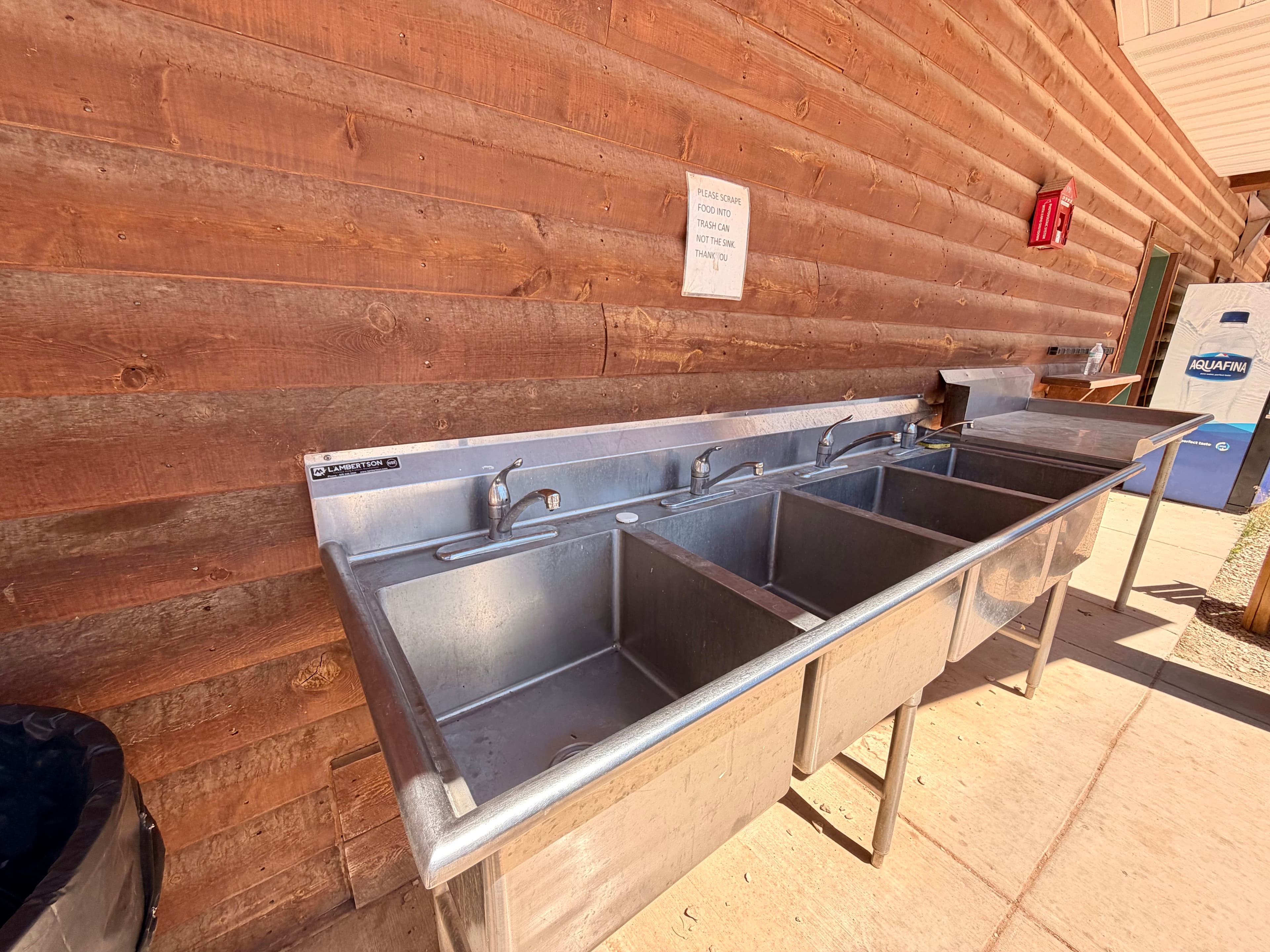 A metal three-compartment sink mounted against a wooden wall.