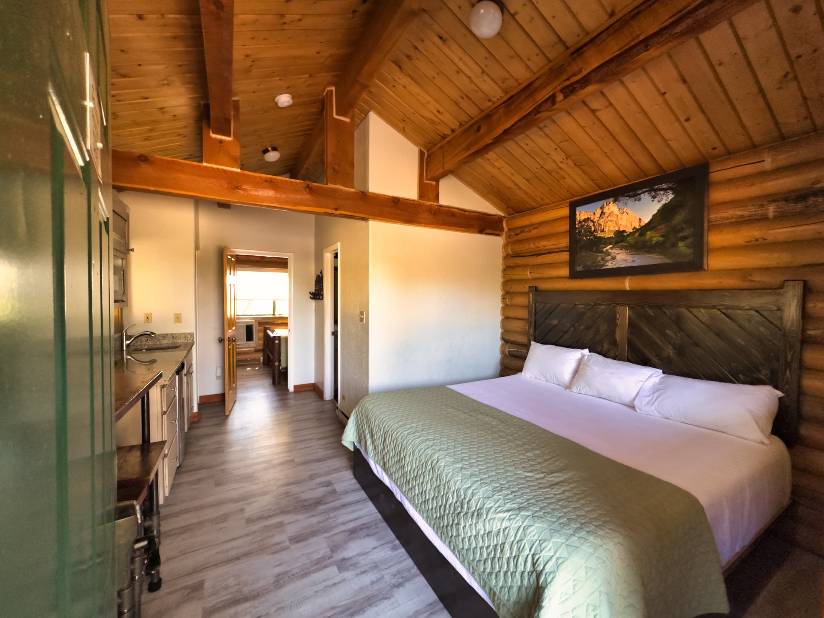 A cozy cabin interior featuring a double bed, wooden beams, and a small kitchenette.