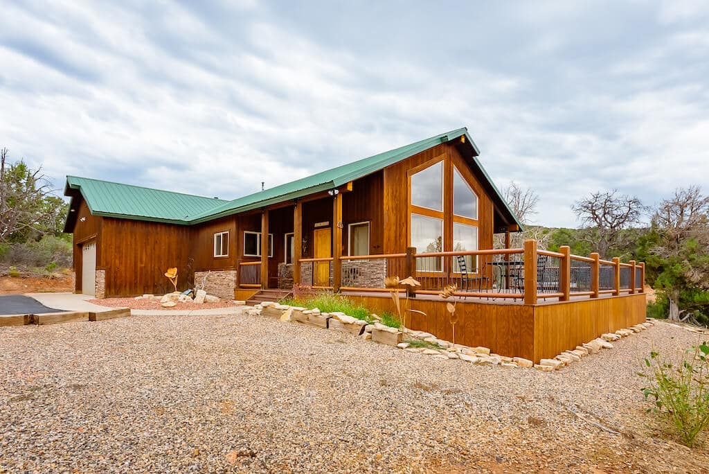A modern wooden house with a green roof and large windows, surrounded by gravel and vegetation.