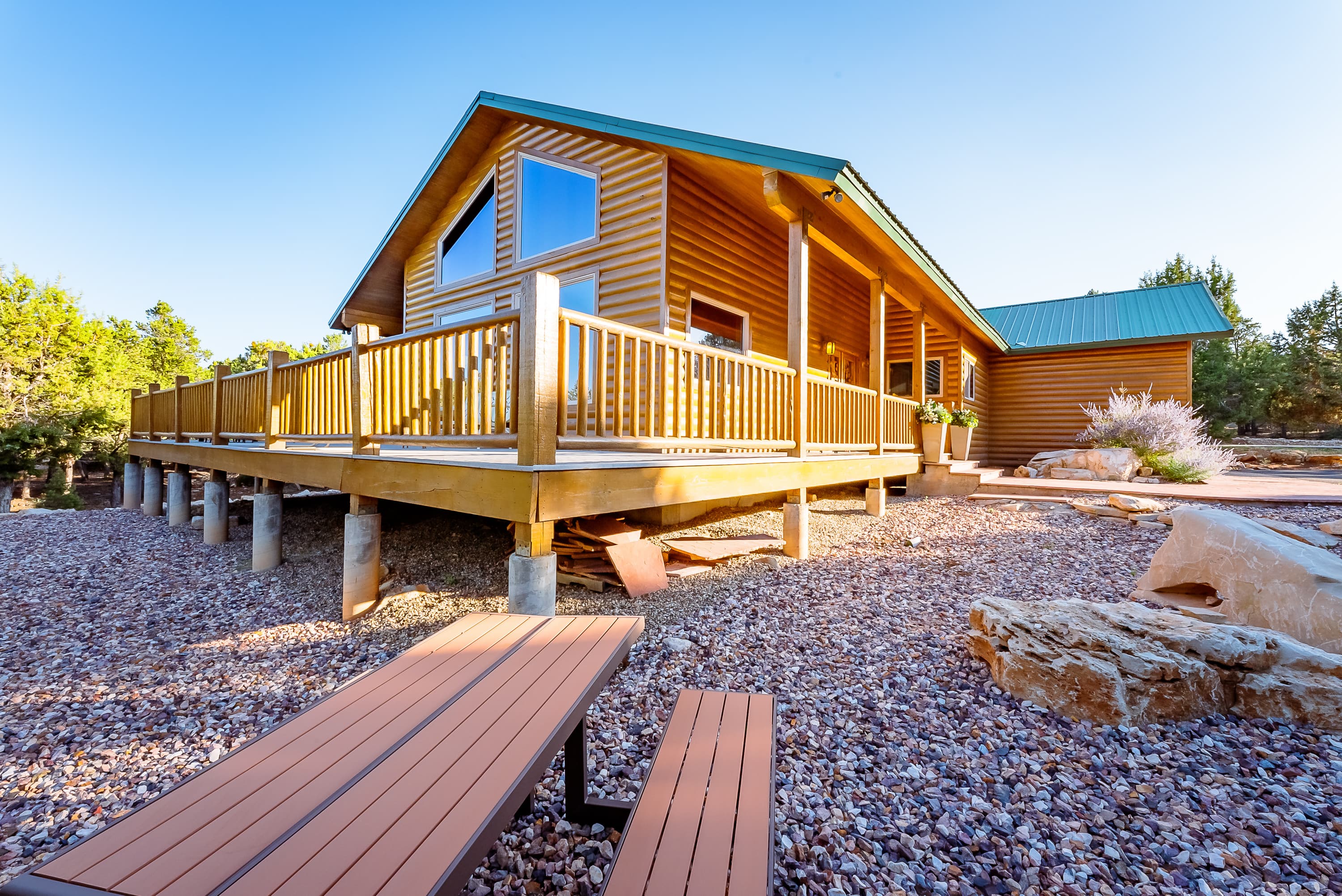 A modern log cabin with a deck, surrounded by rocky landscaping and trees.