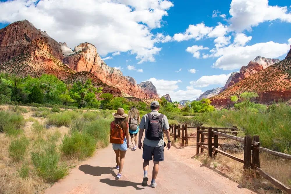 Four hikers walking along a trail with red rock formations and blue skies in the background.