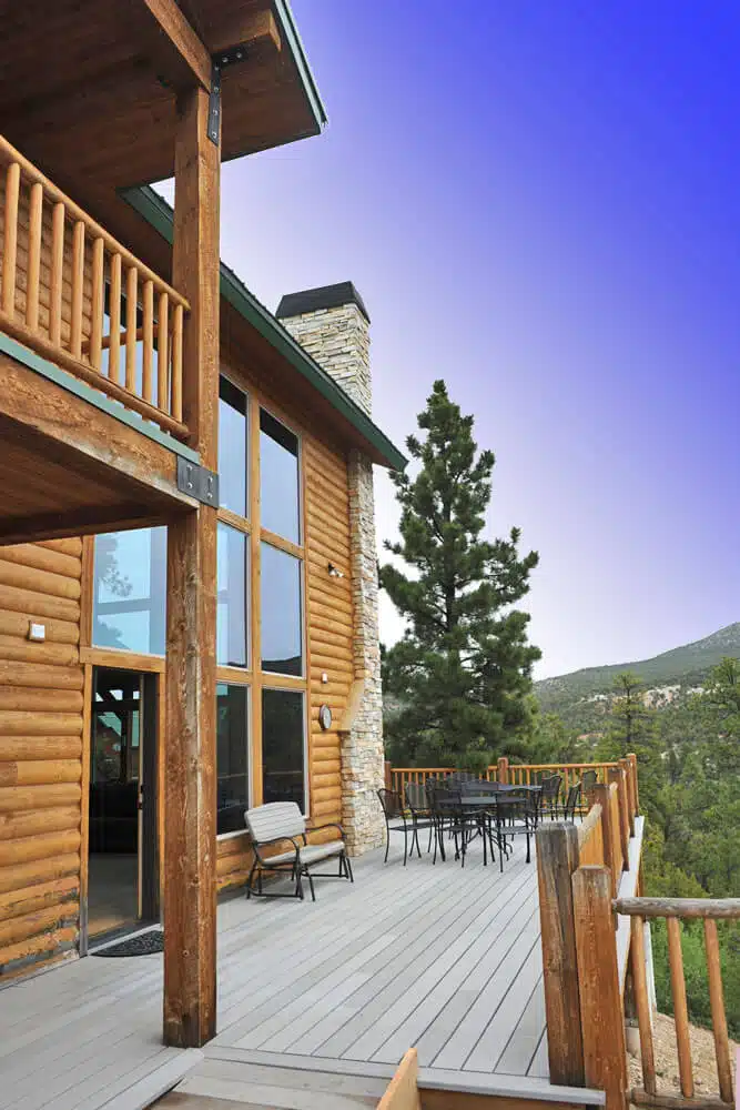A modern log cabin deck overlooks a forested landscape under a clear blue sky.