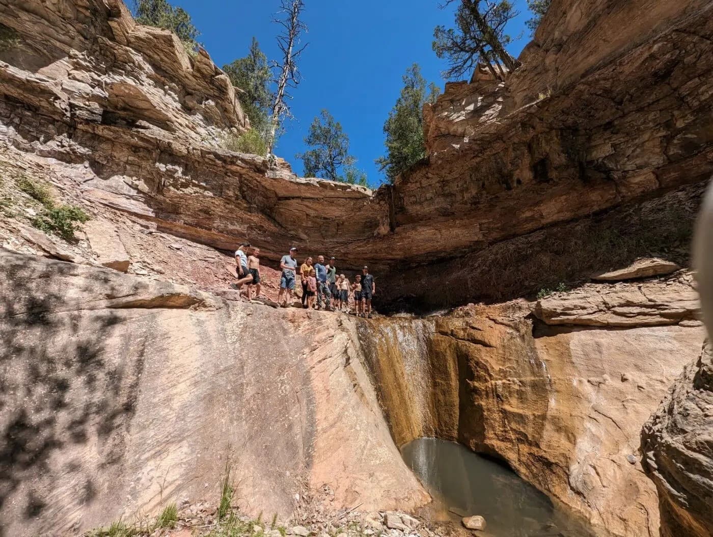A group of hikers stands near a small waterfall in a rocky canyon under a clear blue sky.