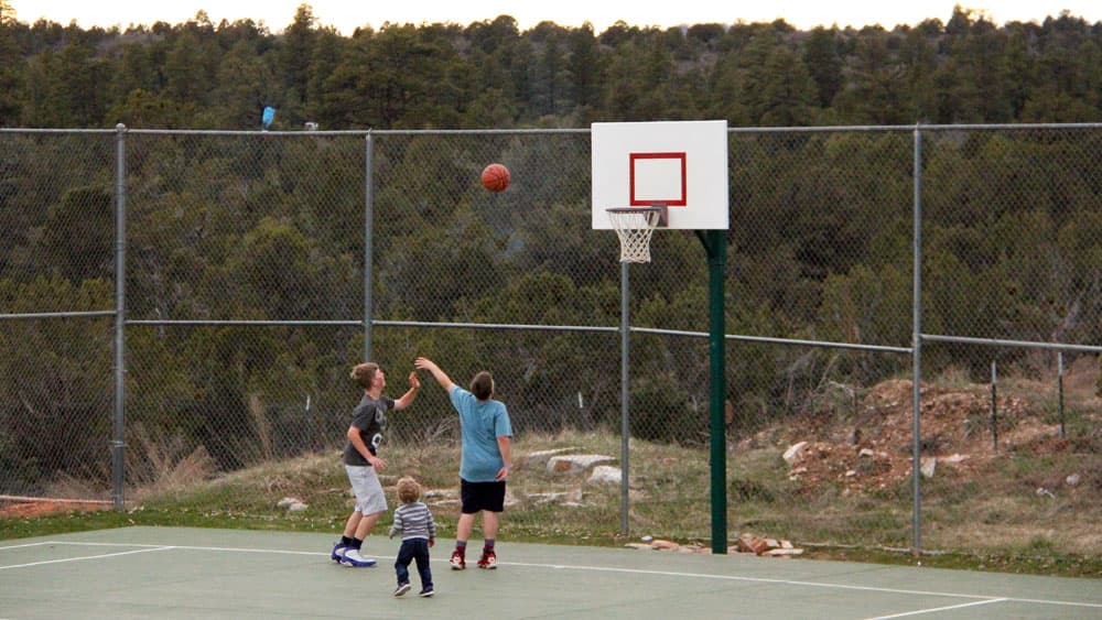 Three children play basketball on an outdoor court surrounded by trees.