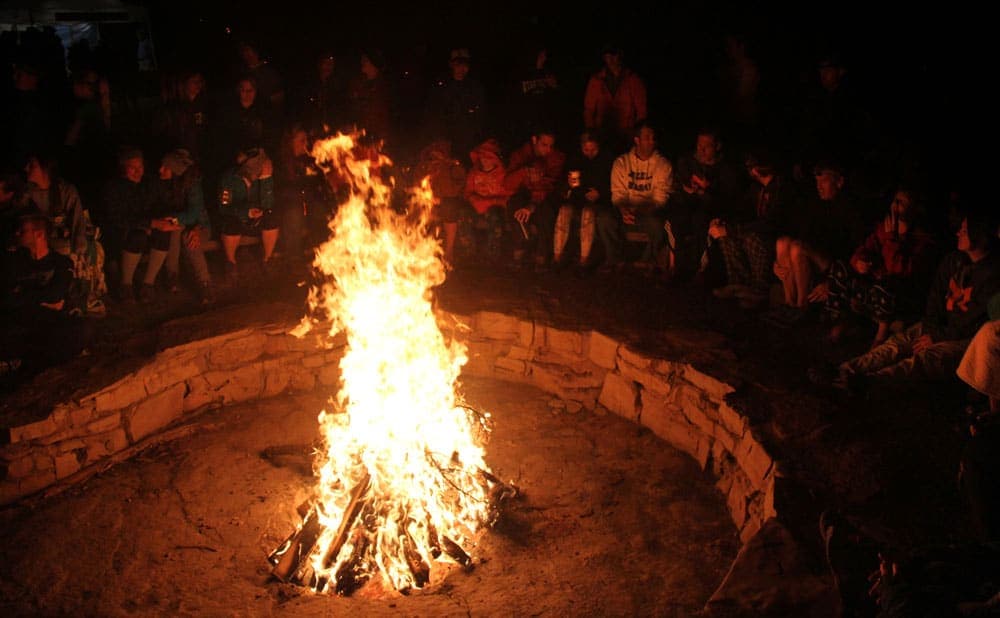 A large campfire surrounded by people sitting in the dark.