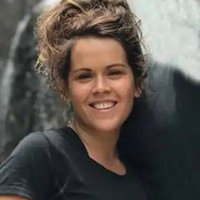 A smiling person with curly hair stands against a dark background.